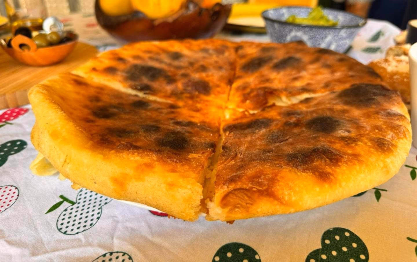 Целое хачапури по-имеретински, румяное и в пузырьках, разрезанное на четыре части на тарелке, за ним оливки, персики и масло
