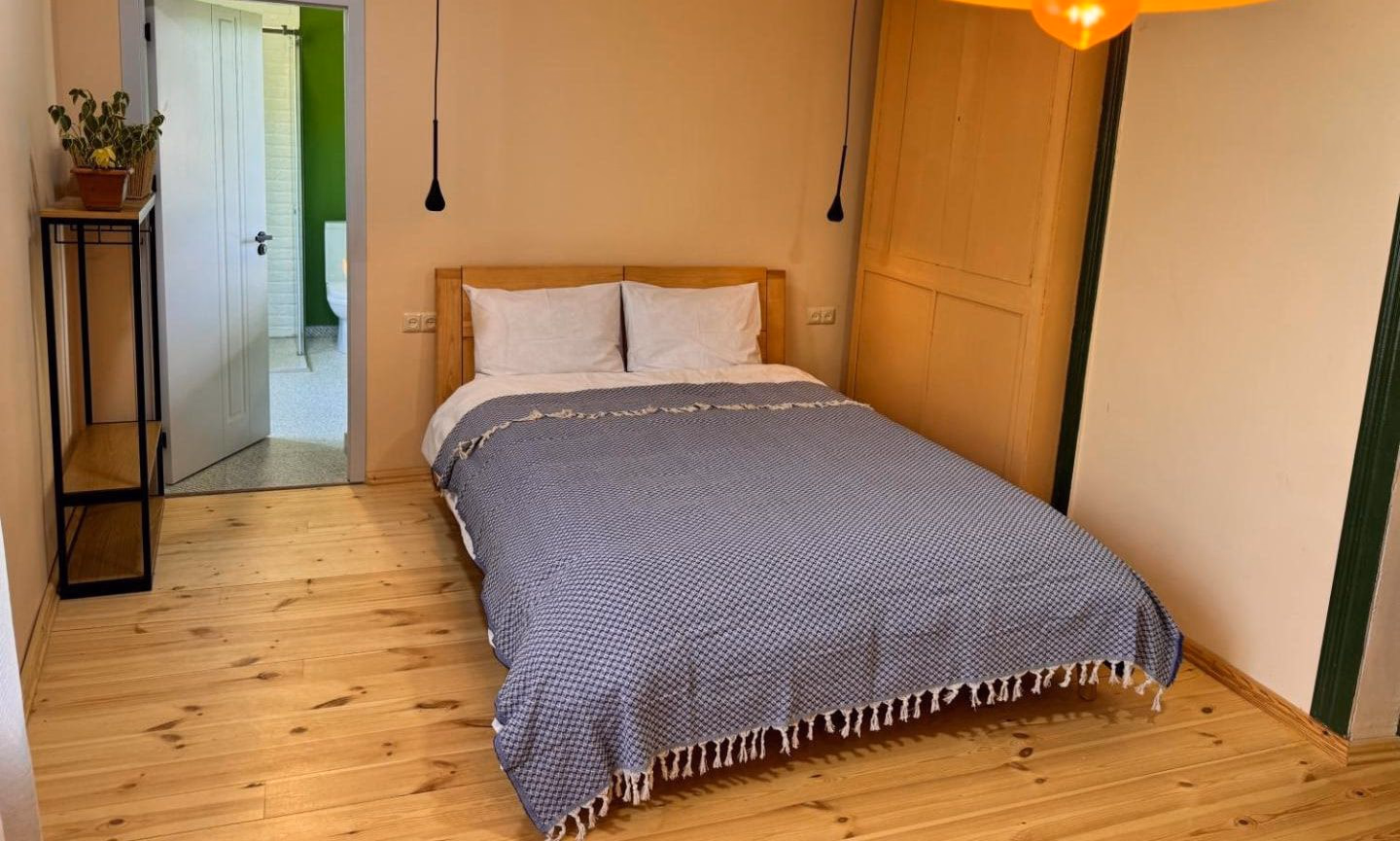 A bedroom with apricot walls and wide pine floorboards, an oak bed with a blue waffle cover, and an open door showing a bright green bathroom